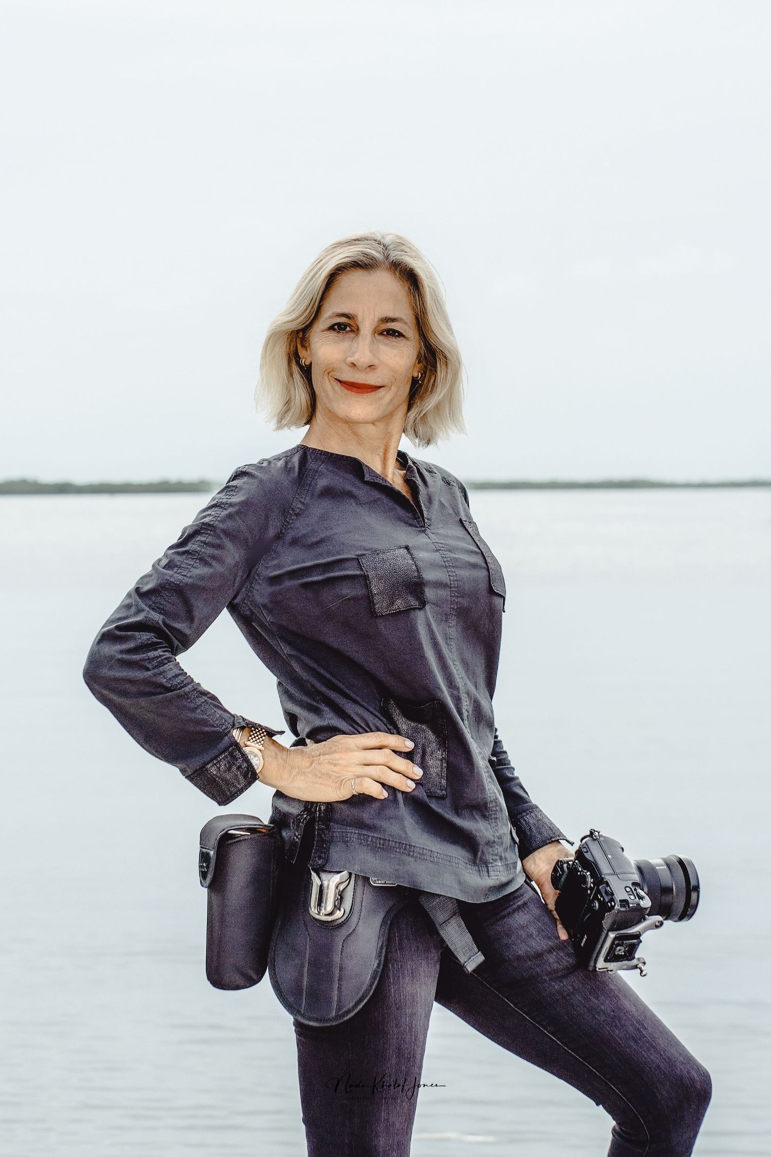 Professional photographer in navy outfit poses with camera gear against water backdrop.