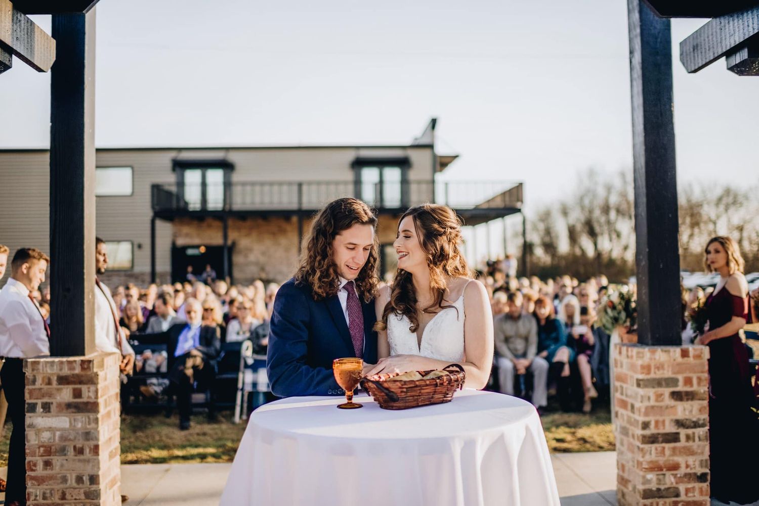 Wedding ceremony features shared moment at small table with guests watching in background.