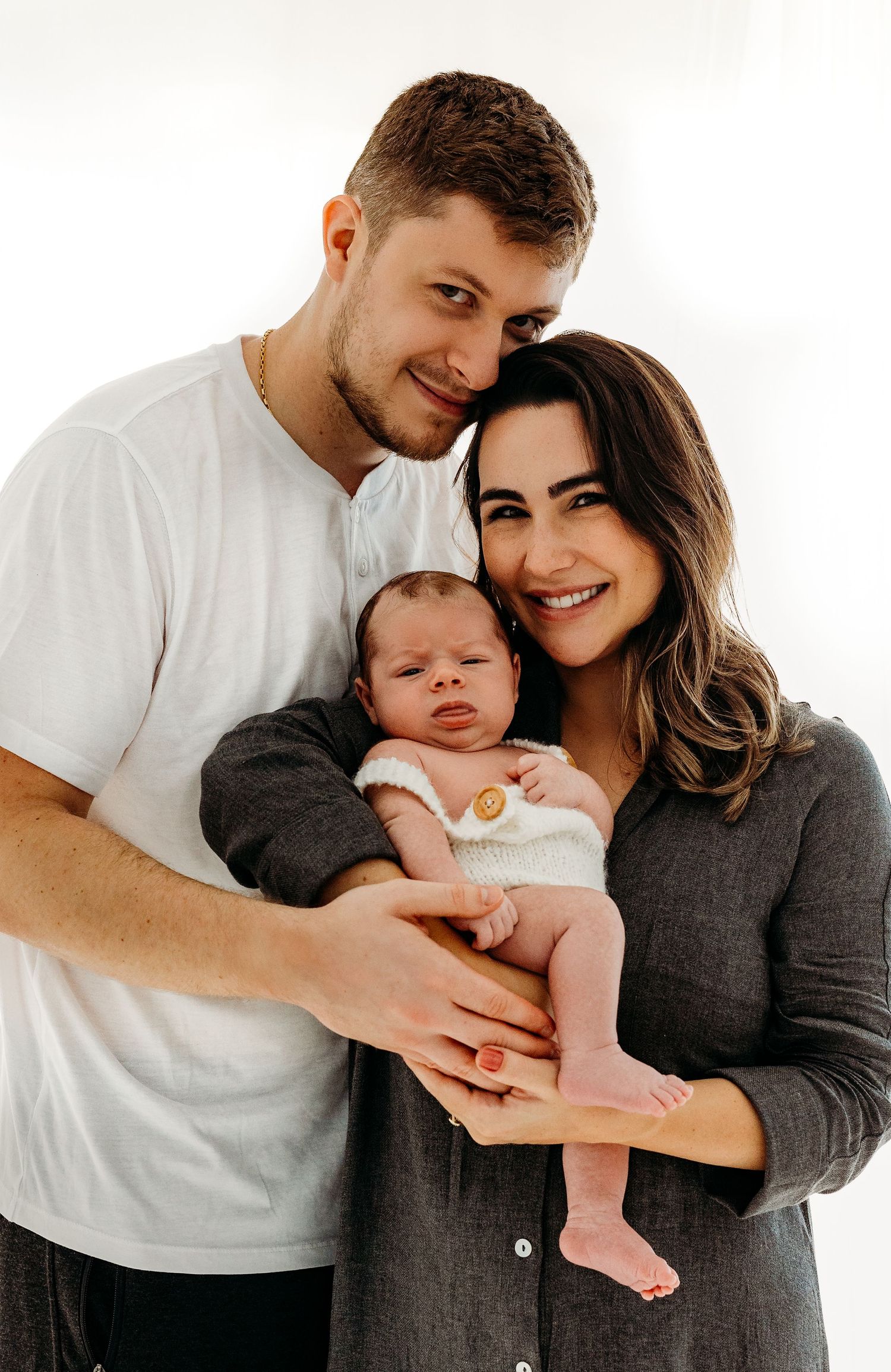 Young couple in casual wear holds their newborn baby against white background.