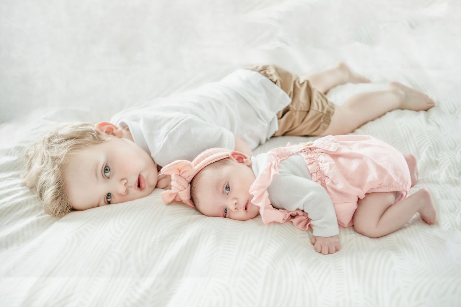 Two young children in light colored clothing lay together on white bedding.