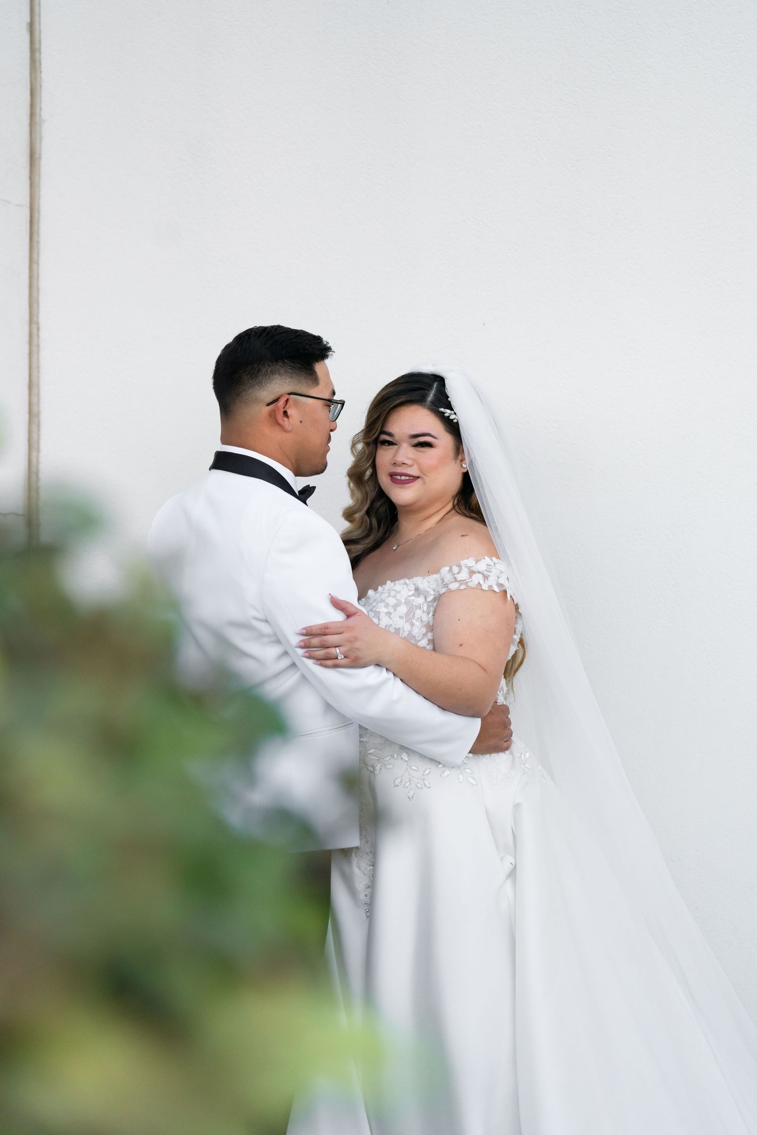 A bride and groom share an intimate moment against a white wall with greenery in the foreground.