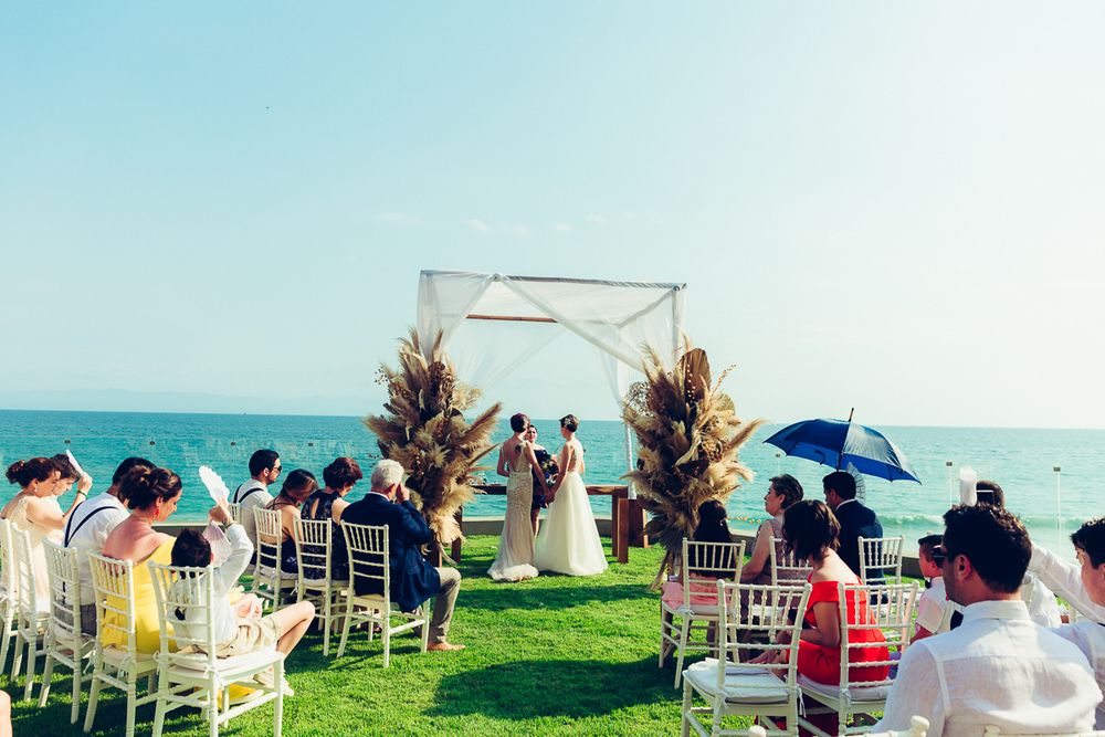 An oceanfront wedding ceremony with white chairs and guests holding parasols under a bright blue sky.