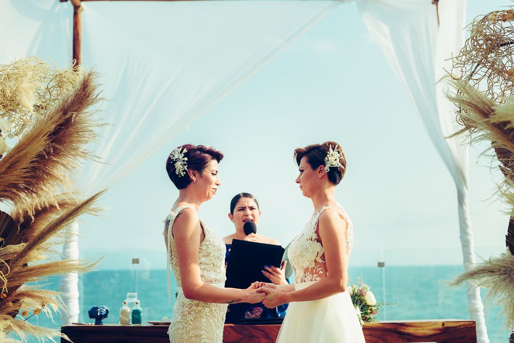 A beachside wedding ceremony under a white arch decorated with pampas grass against the ocean backdrop.
