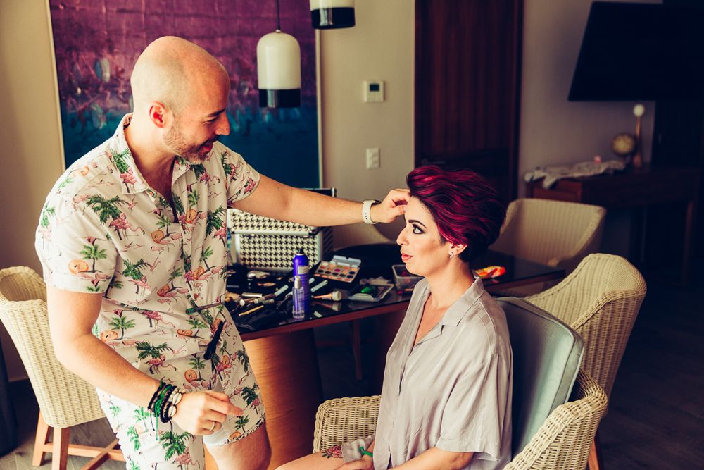 A make-up artist in a floral shirt applies cosmetics to a client seated in a chair during wedding preparations.