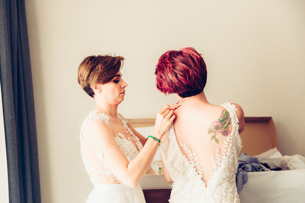 Two people in white lace wedding dresses helping each other get ready.