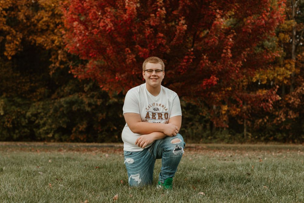 Person kneeling on grass wearing white t-shirt and ripped jeans with red autumn foliage behind.