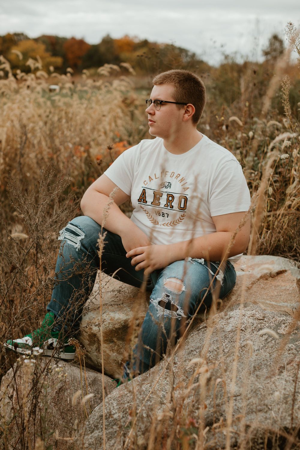 Person in white t-shirt and ripped jeans sitting on rock in autumn field.