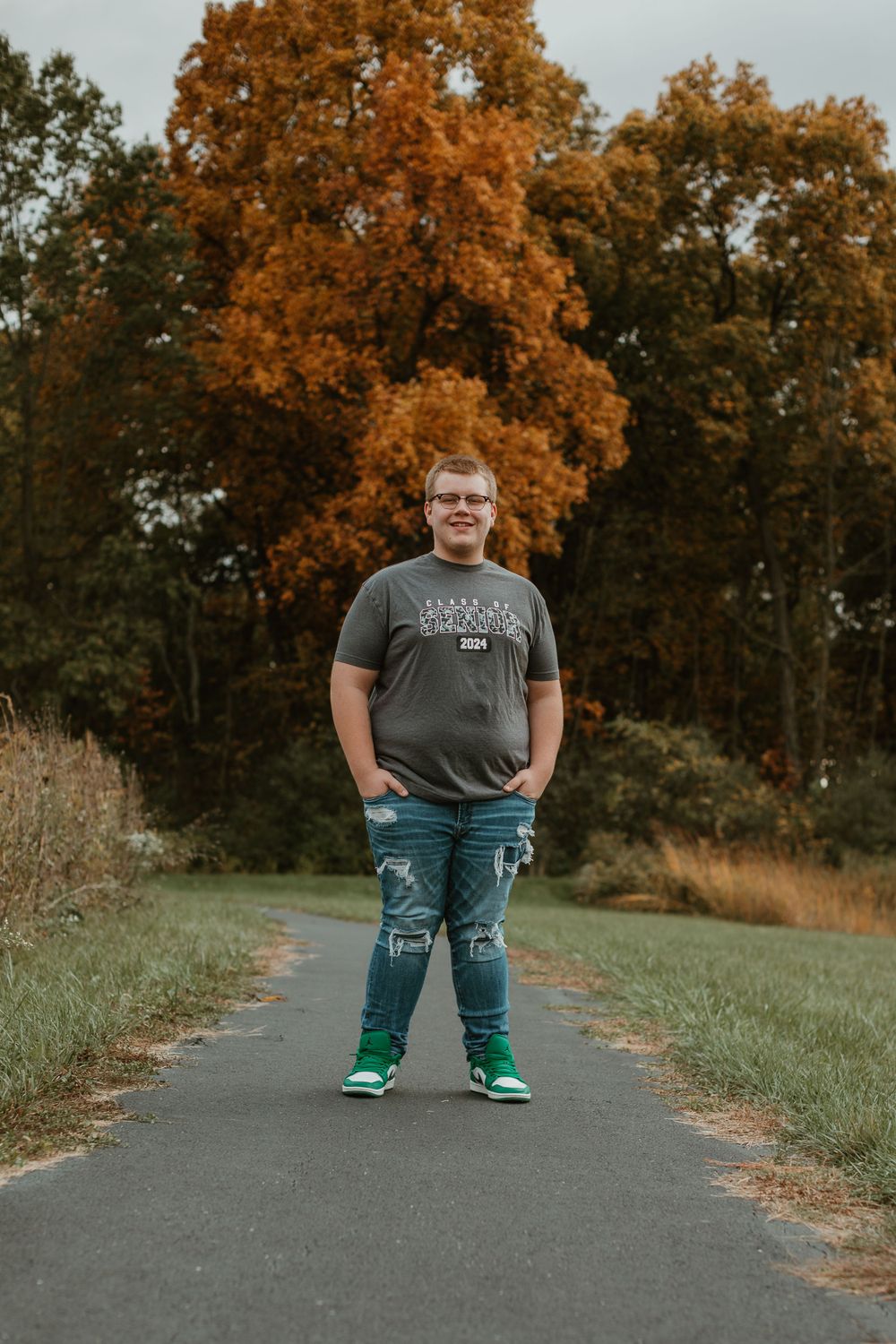 Person in grey t-shirt and ripped jeans standing on path with fall foliage in background.
