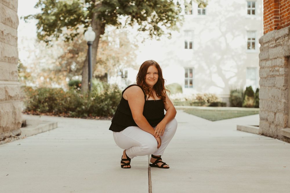 Person in black top and white pants crouching on concrete path near building.
