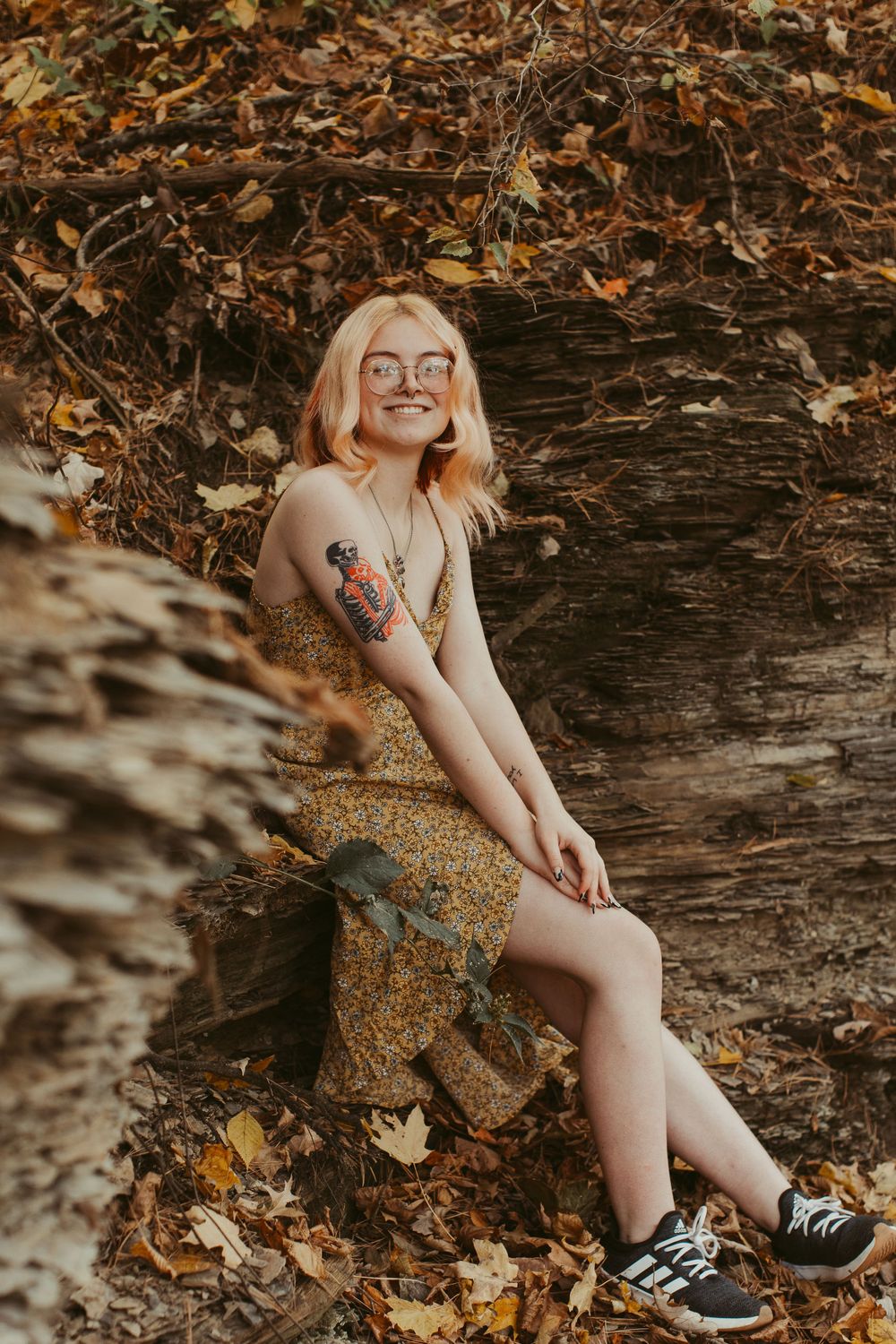 Portrait of someone in floral dress with tattoos sitting on rocky ledge surrounded by autumn leaves.