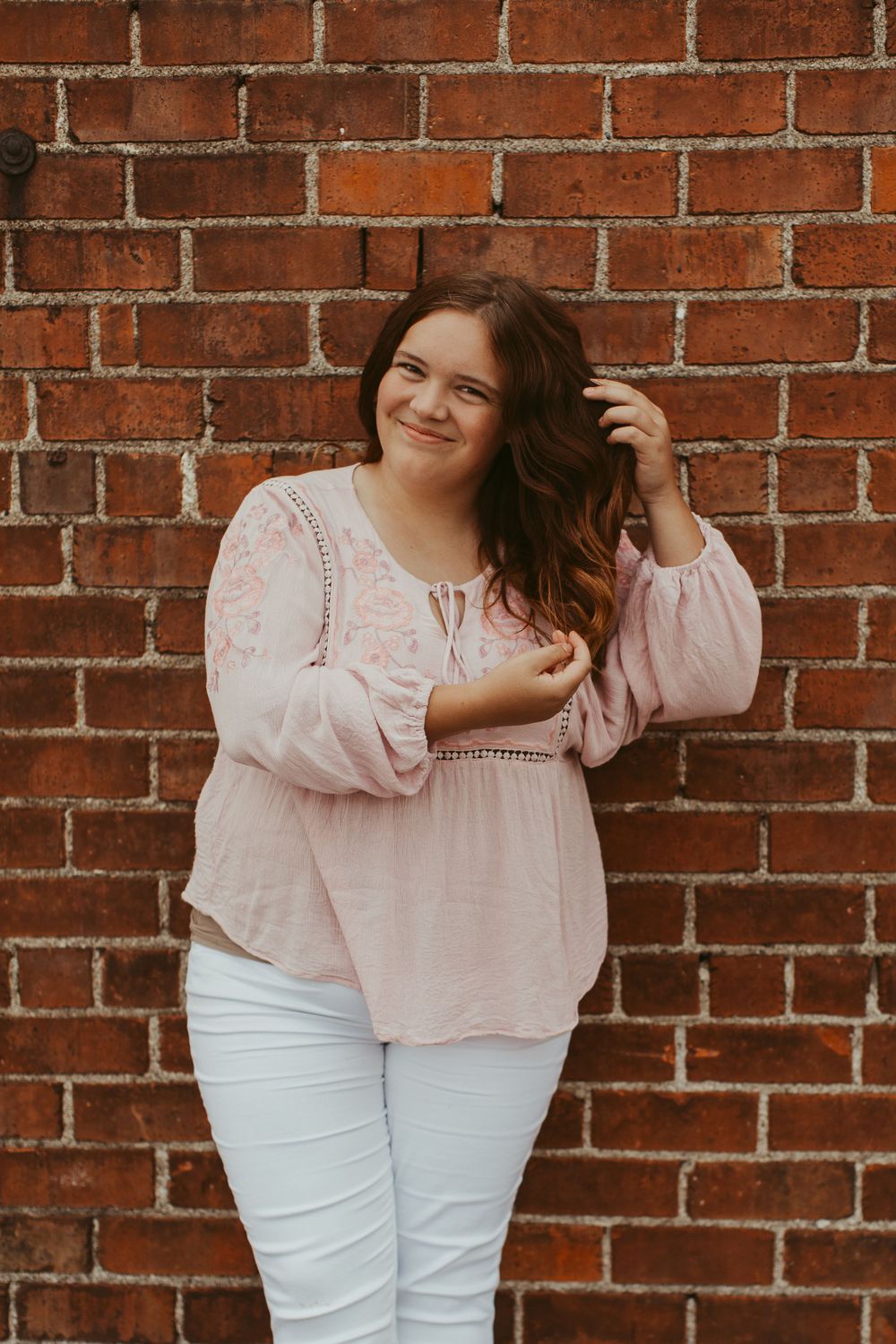 Person in pink peasant blouse and white pants posing against brick wall.