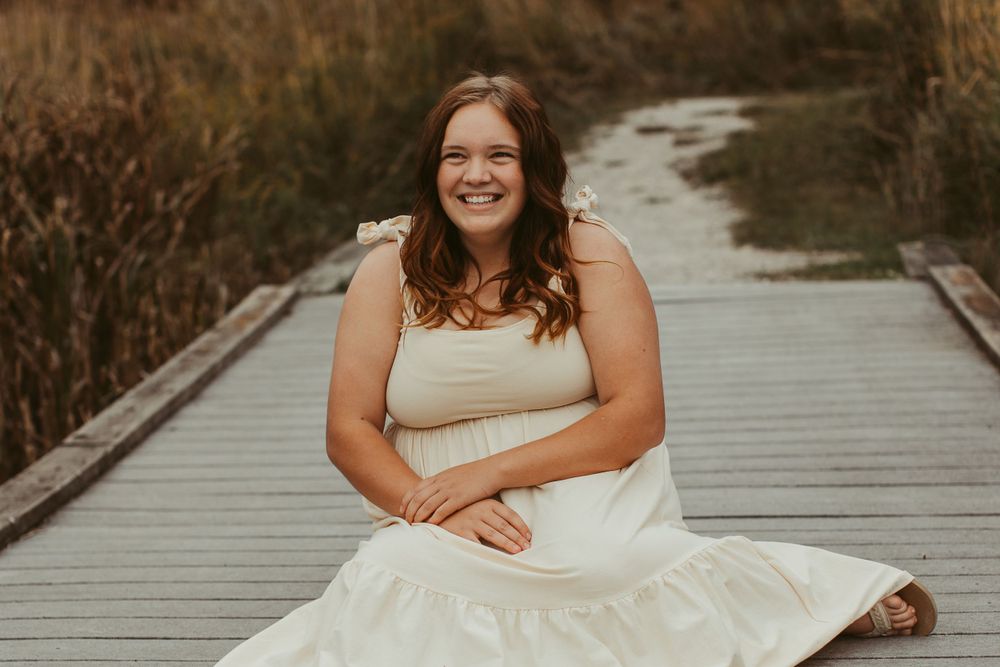 Person in white dress sitting on wooden boardwalk surrounded by autumn grass.