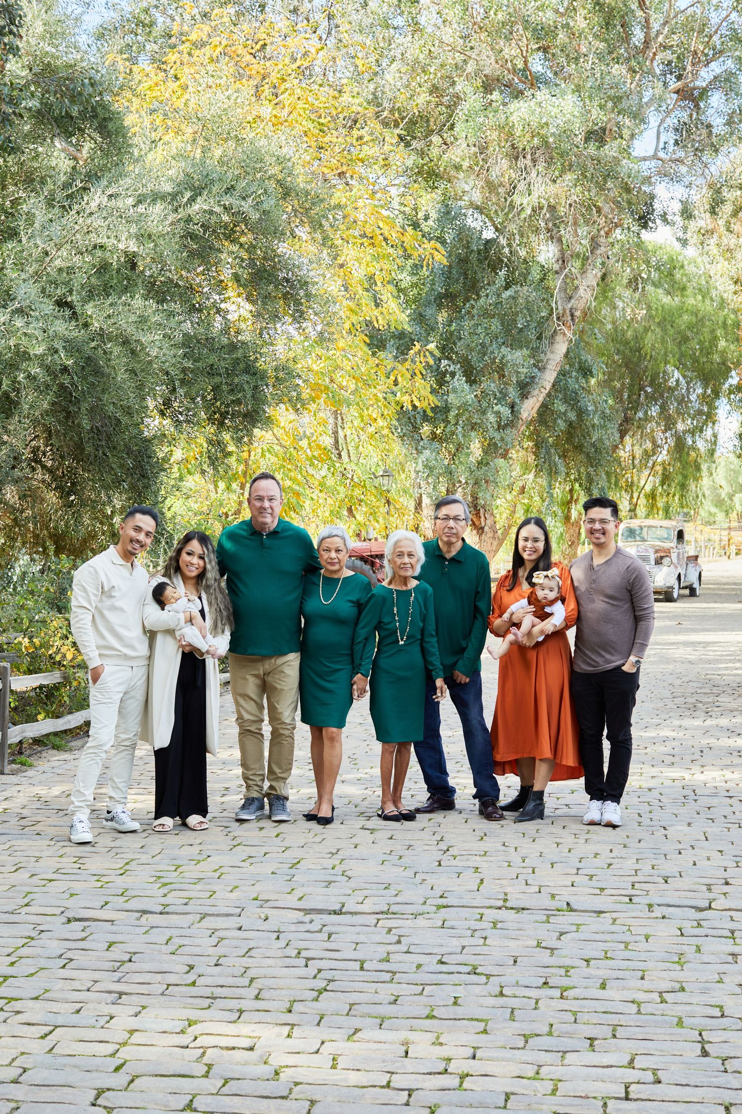 Extended family in coordinated outfits poses together on cobblestone path surrounded by trees.