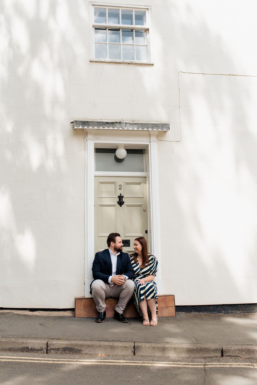 A couple sits intimately on a doorstep of a white building with shadows cast from trees.