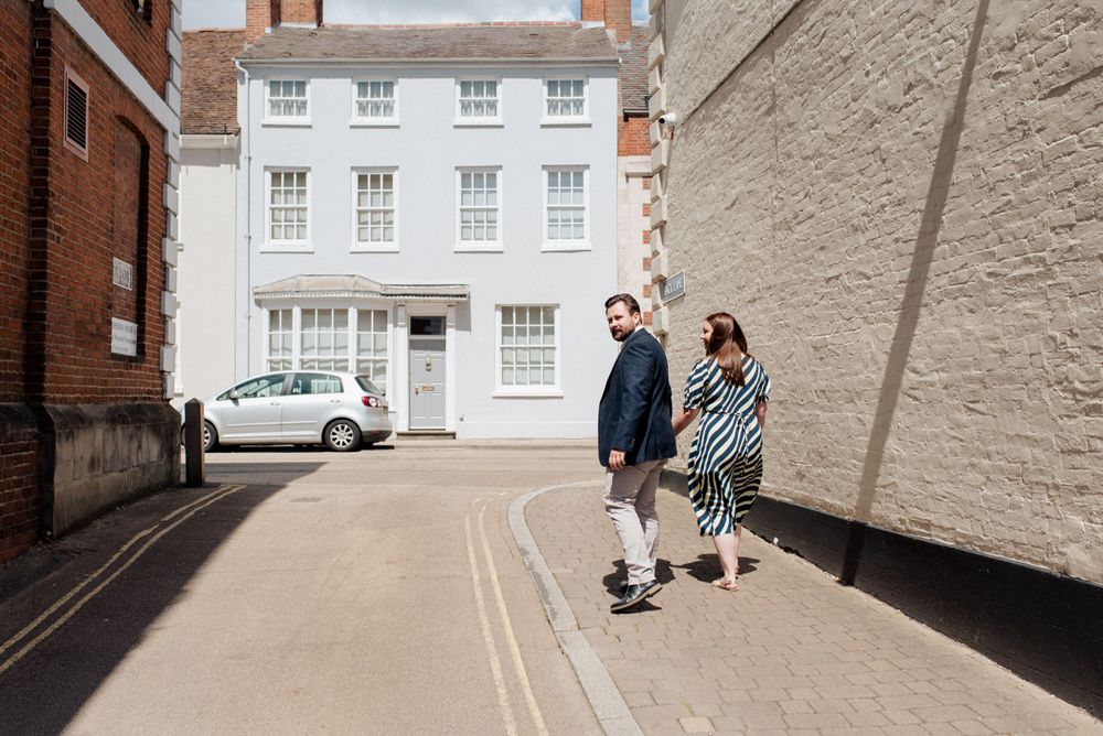 A couple walks down a quaint street with classic British architecture in the background.
