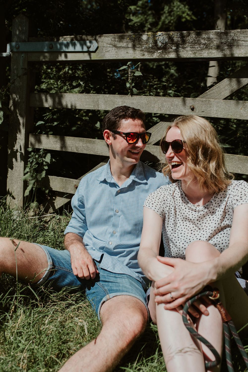 Couple sitting on grass laughing together on sunny summer day wearing sunglasses.