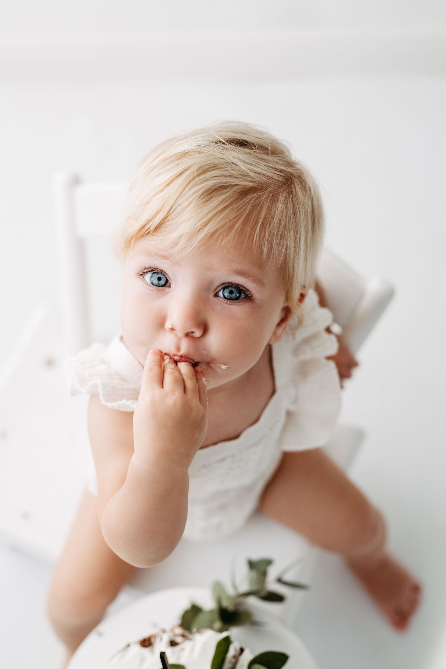 Sweet portrait of toddler in white dress with finger in mouth.