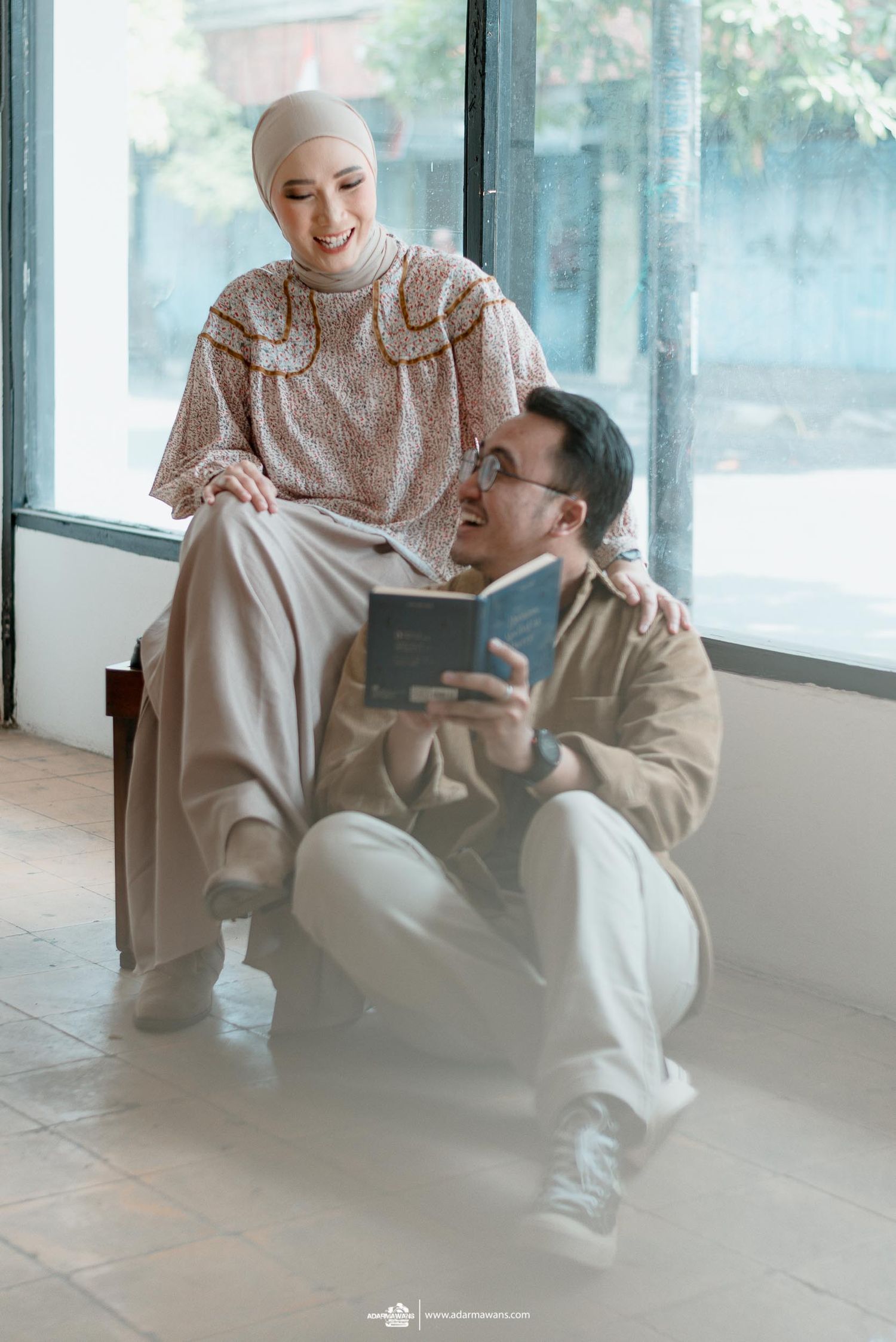 A couple in neutral tones shares an intimate moment reading together by a sunny window.