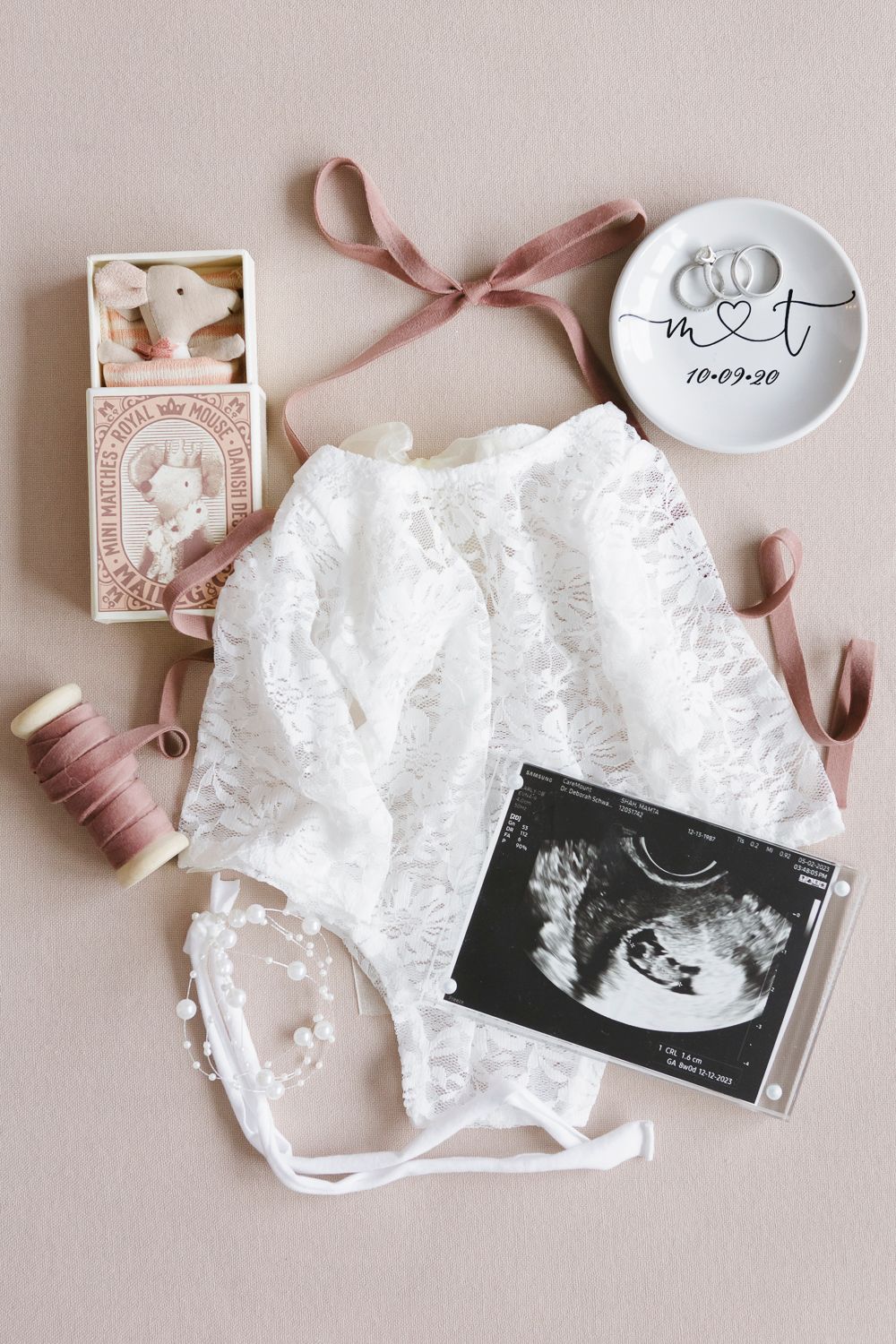 Flatlay of sonogram with dress, bow, toy, and wedding rings, photographed during a newborn session at home by Paula O