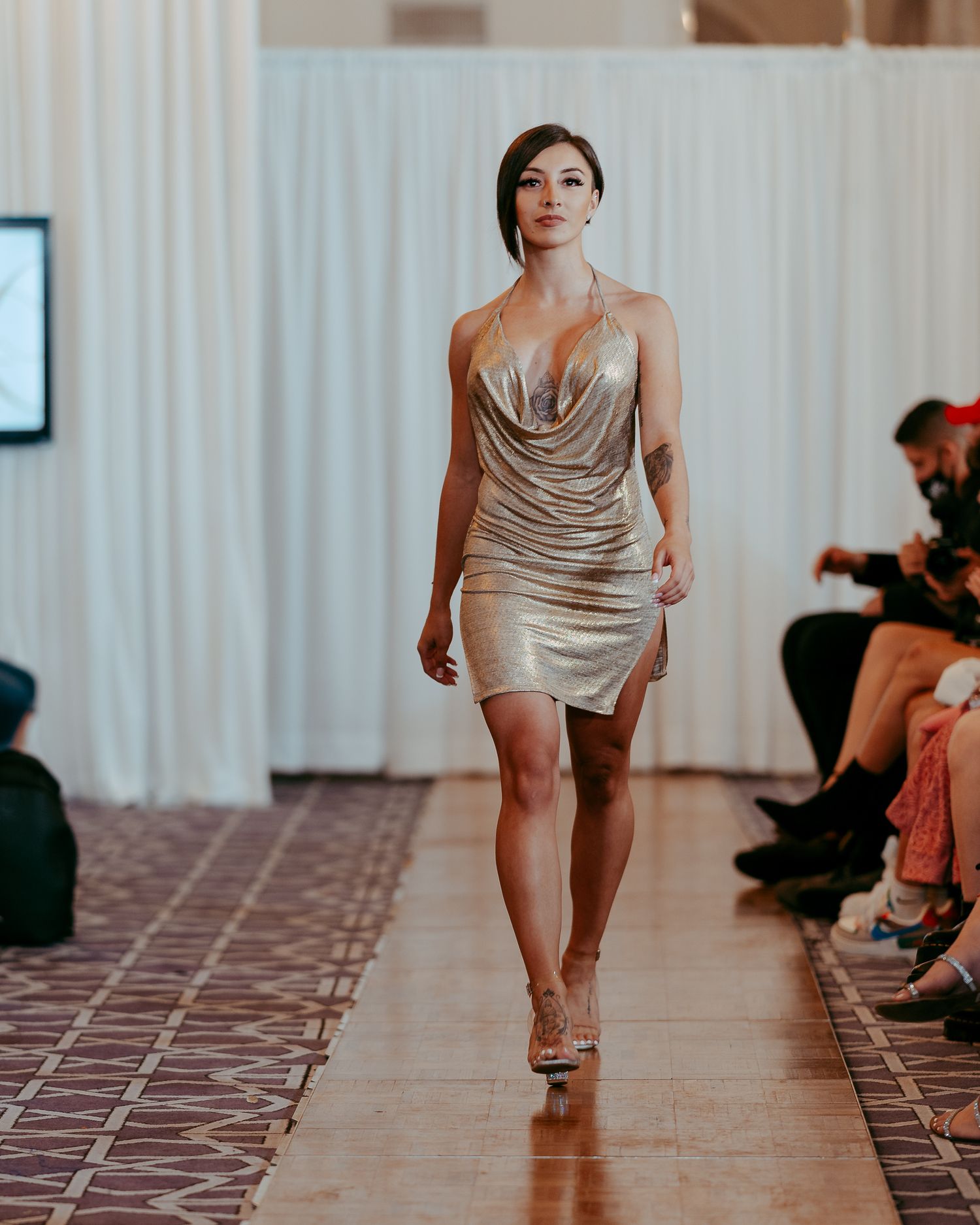 Model in flowing beige mini dress walks down tiled runway at indoor fashion show.
