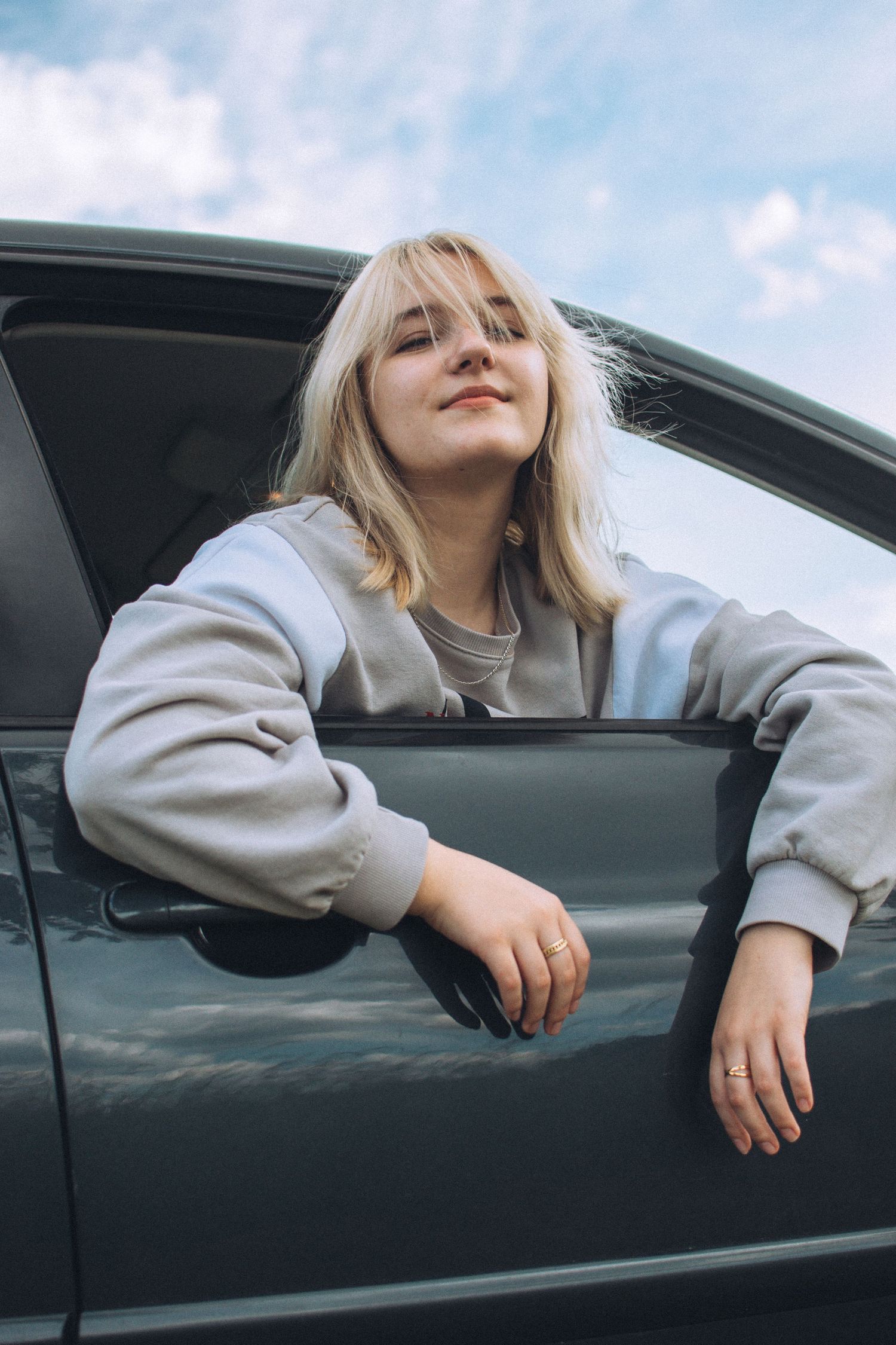 Person in gray sweatshirt leaning out of car window against blue sky backdrop.