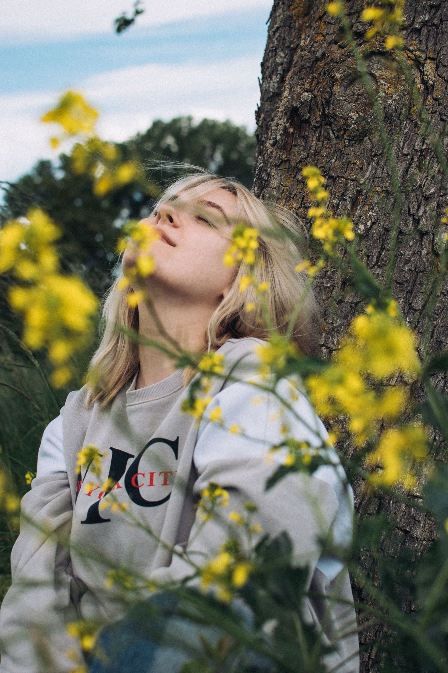 Person in gray sweatshirt resting against tree trunk surrounded by yellow flowers.
