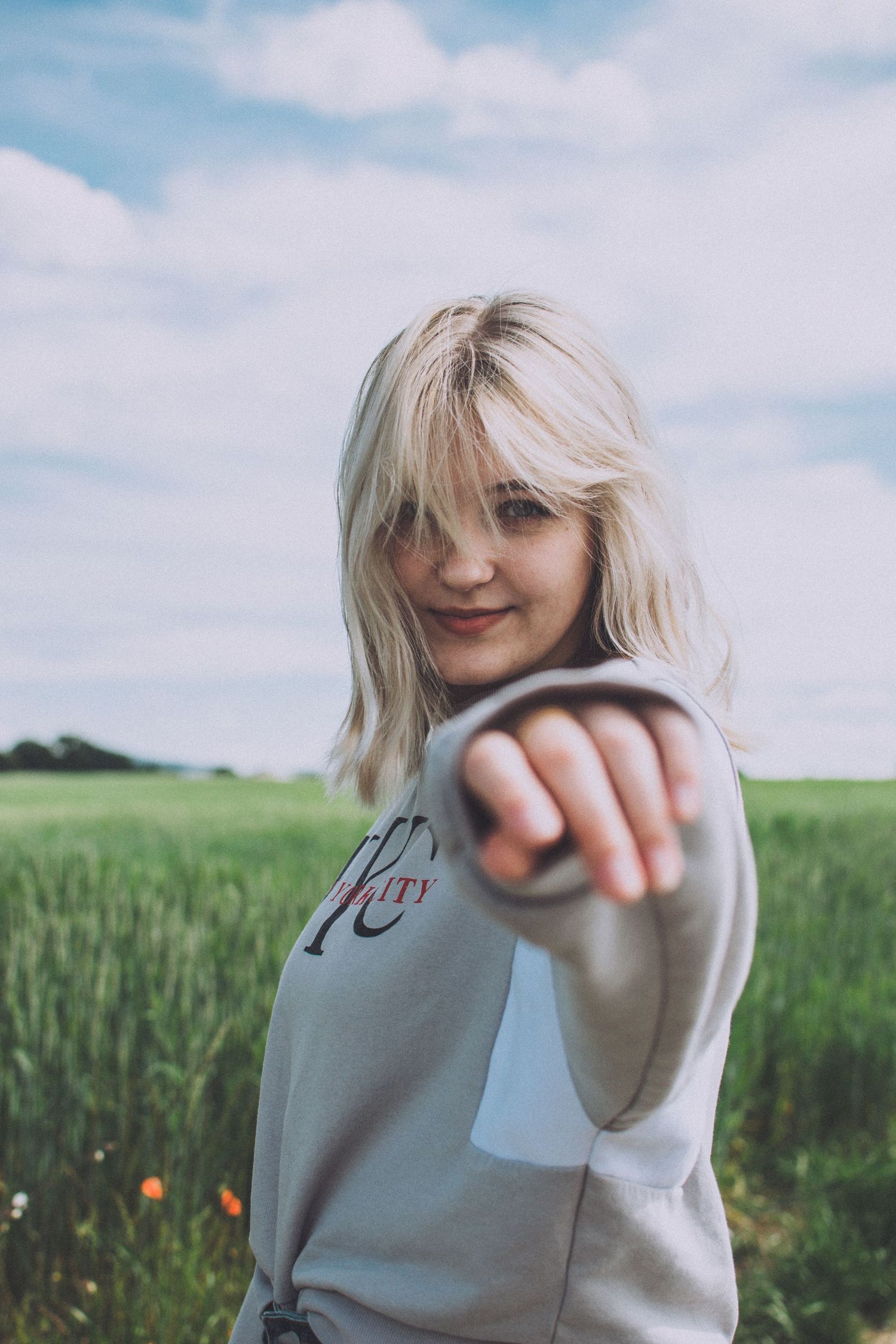 Person in gray sweatshirt pointing at camera while standing in green field.