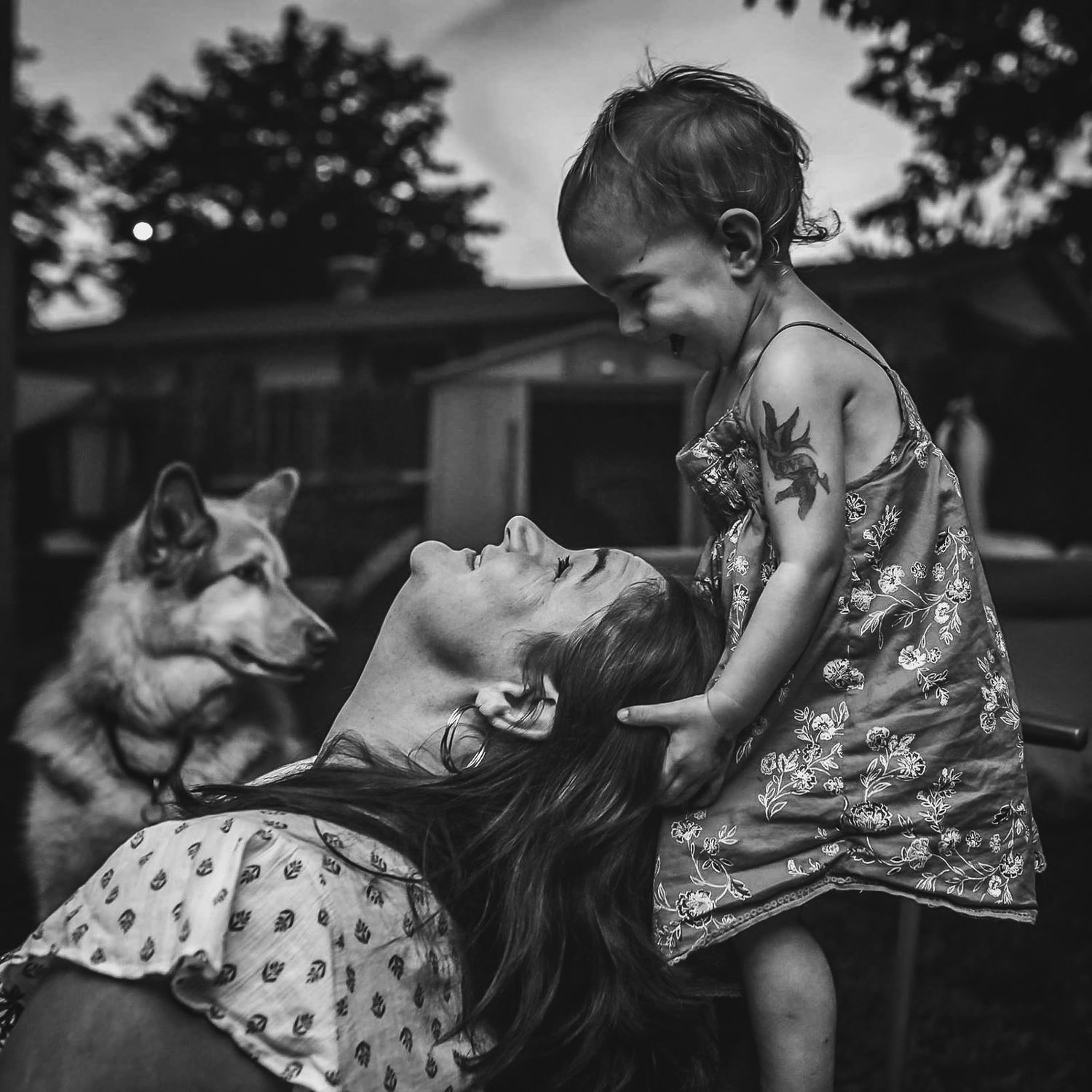 BW image of a toddler looking down at her mother as she looks up at her from the ground. A beautiful dog is in the back.