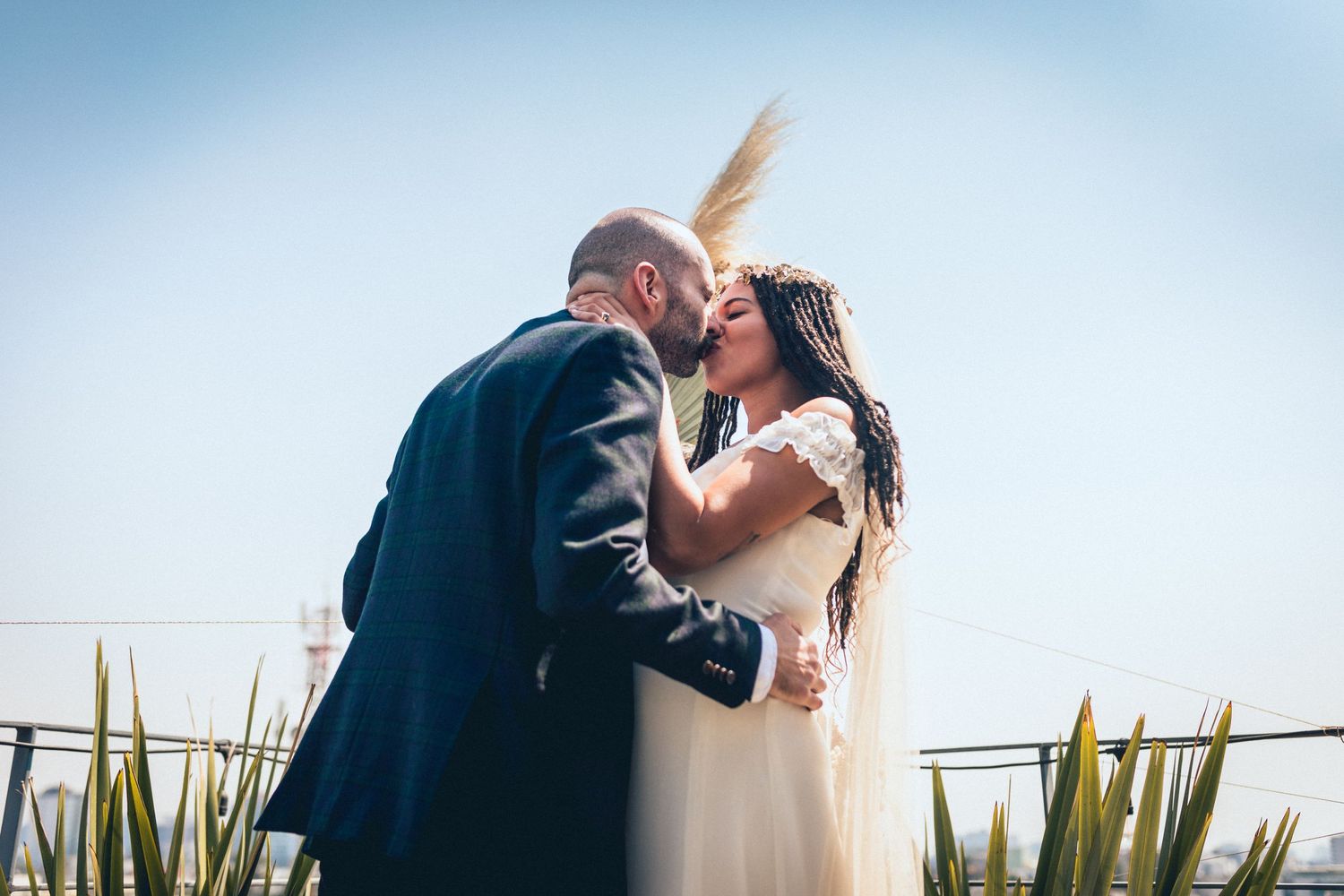 A romantic wedding couple kisses outdoors on a rooftop with blue sky and greenery in the background.