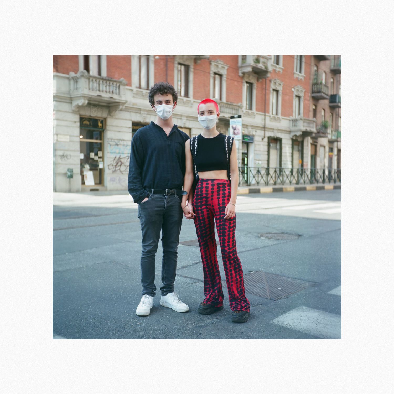 Two people standing on a street wearing contrasting outfits - black sweater and plaid pants against historic building.