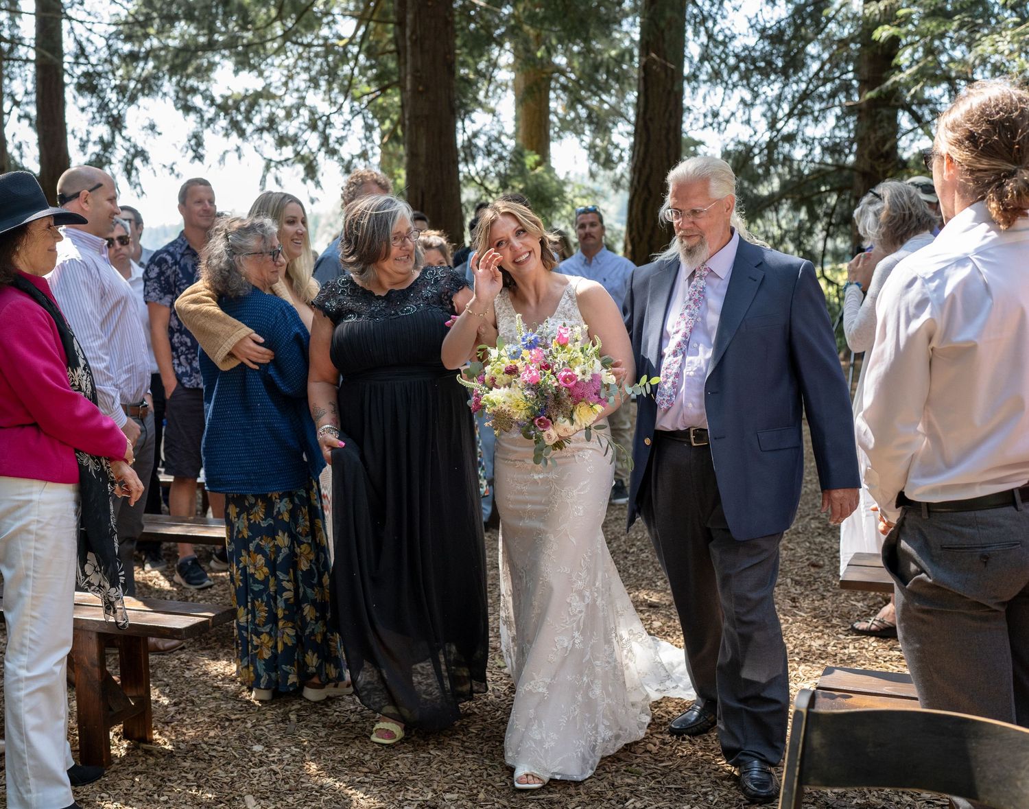 Wedding guests gather around the newlyweds during an outdoor forest ceremony.