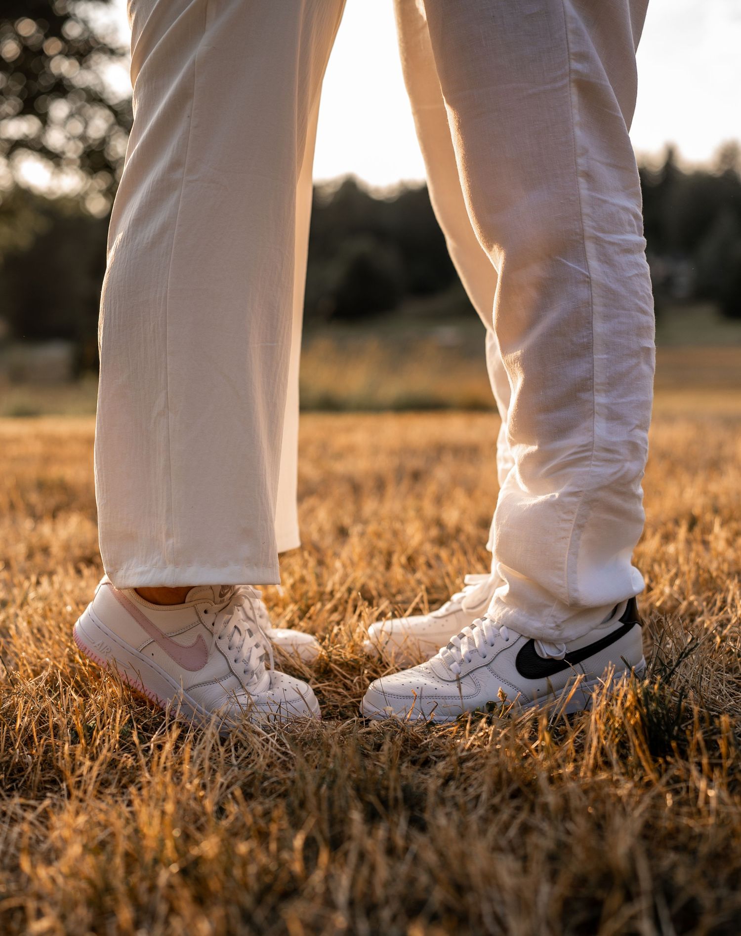 Two pairs of white Nike sneakers standing together in a golden wheat field at sunset.
