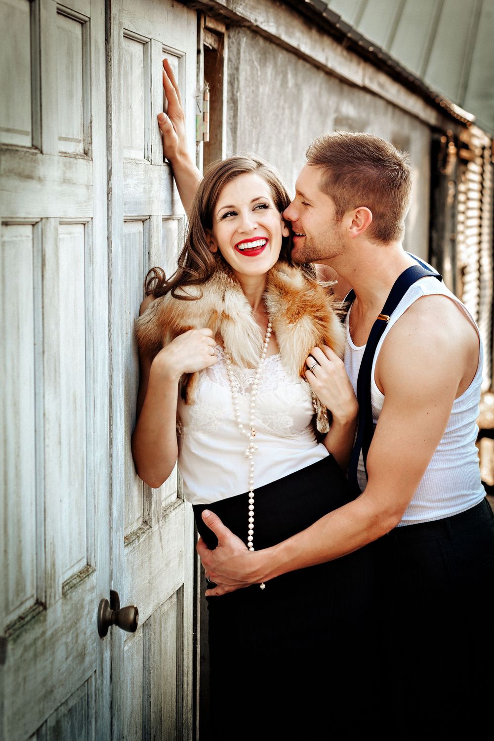 Romantic vintage-inspired couple sharing an intimate moment against a rustic door with fur stole detail and formal attire.