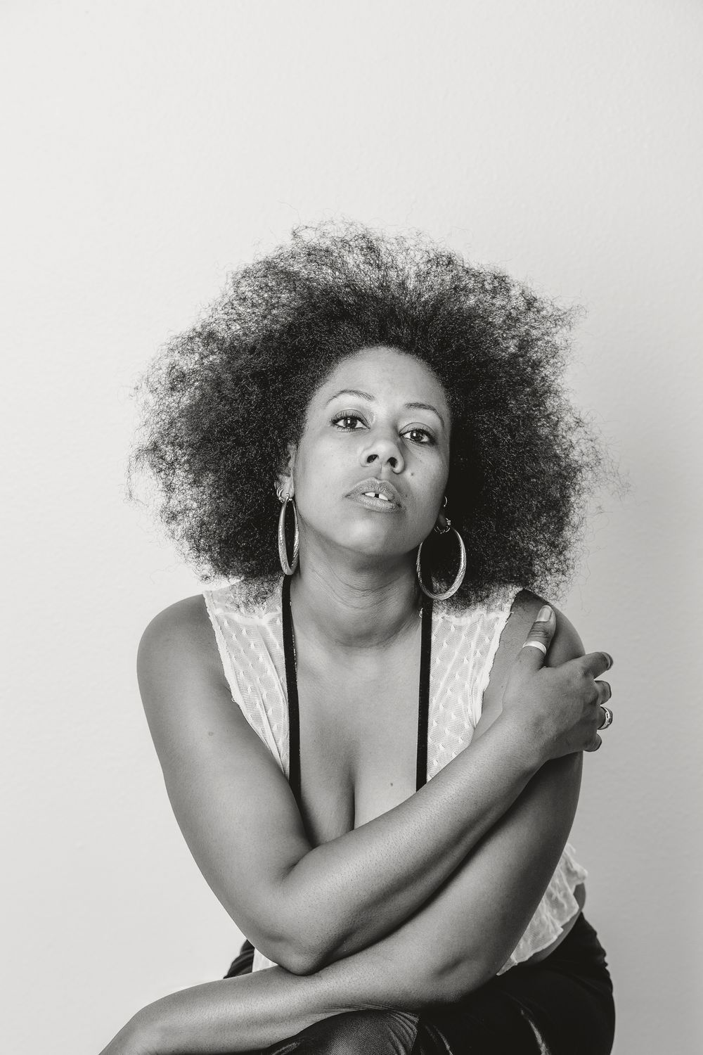 Portrait of woman with natural afro hairstyle wearing hoop earrings and white lace top against neutral background.