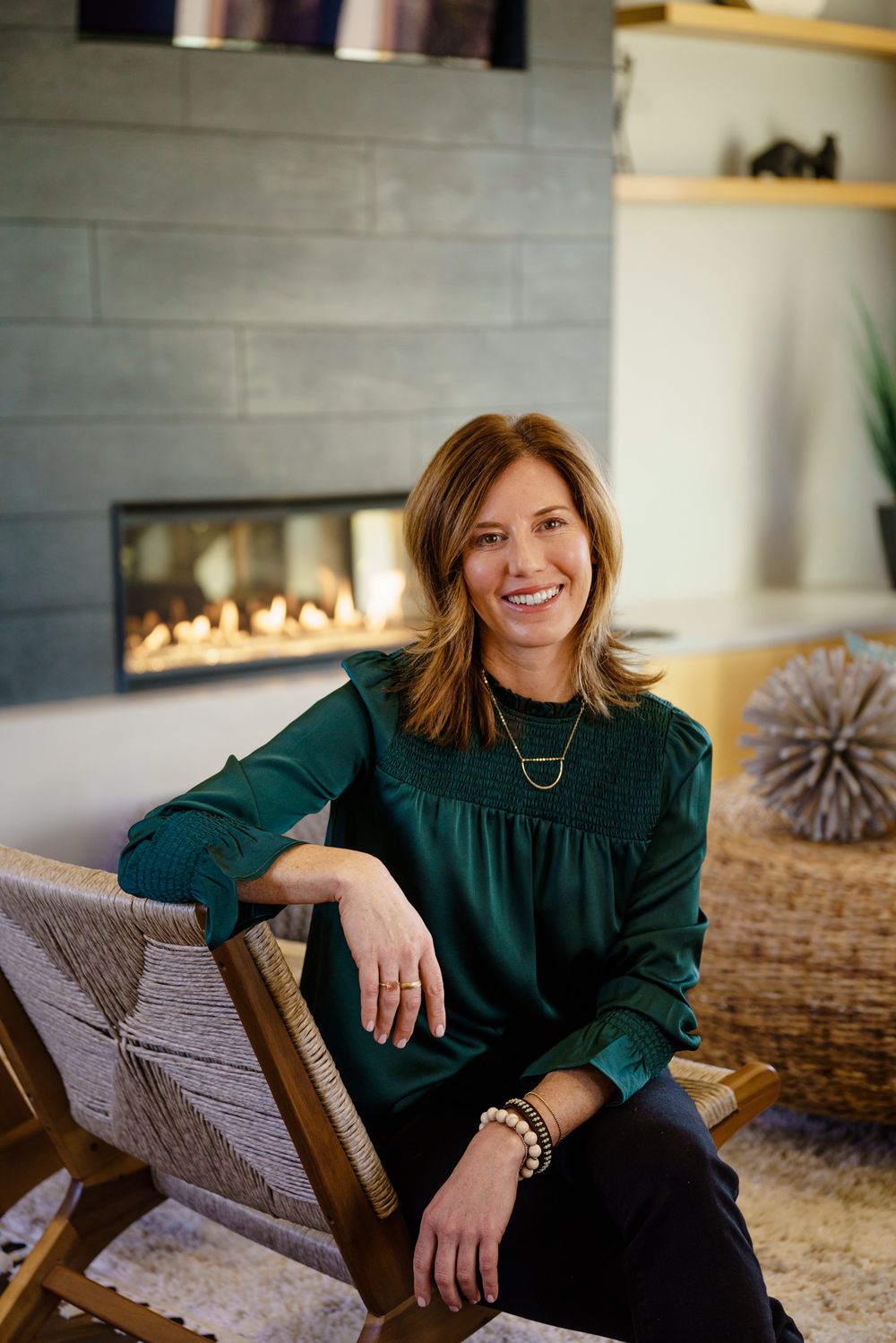 Woman in green sweater sitting in chair by modern fireplace in cozy home setting.