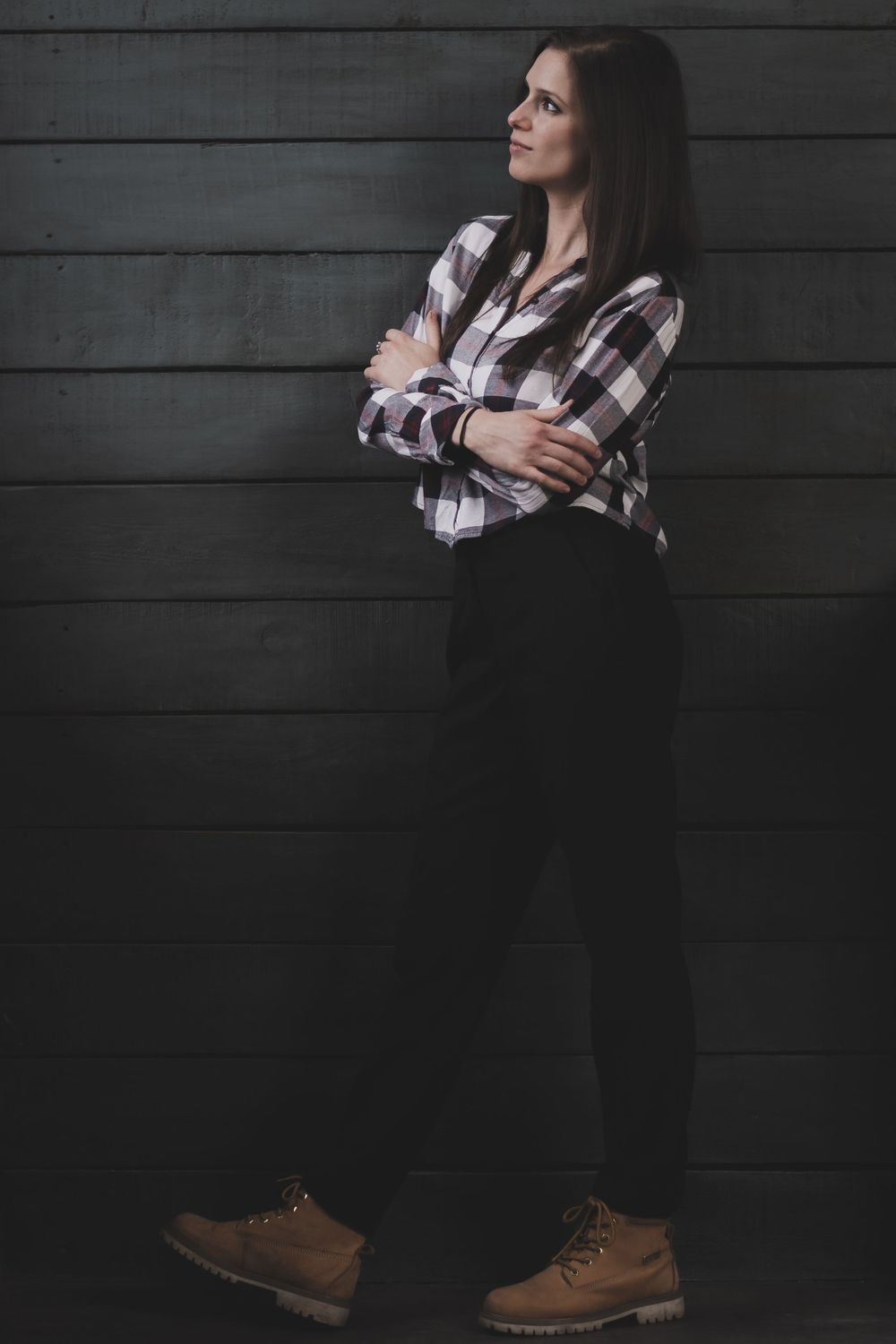 Person in plaid shirt and dark pants standing against gray wooden wall background.