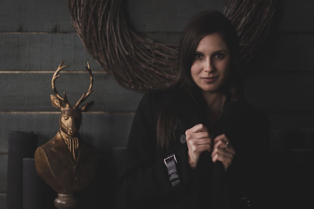 Dark atmospheric portrait with winter clothing and decorative deer statue in background.