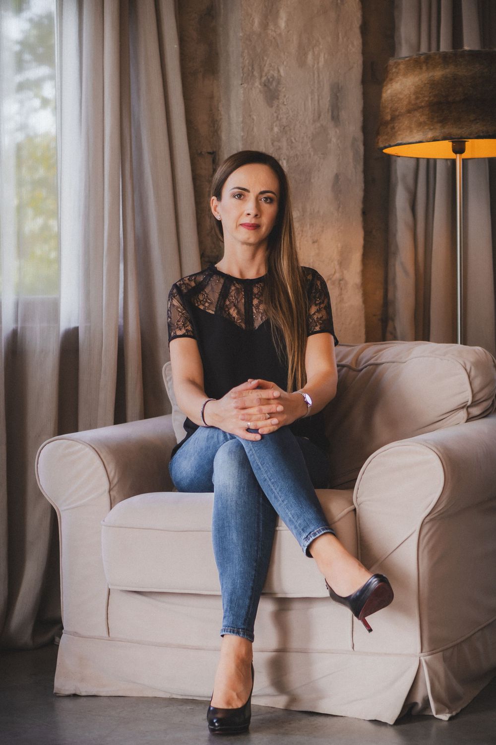 Elegant woman in lace top and jeans sitting poised on white armchair.