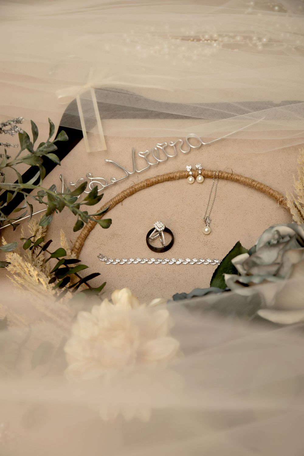 Wedding accessories including rings and jewelry arranged with greenery on beige surface.