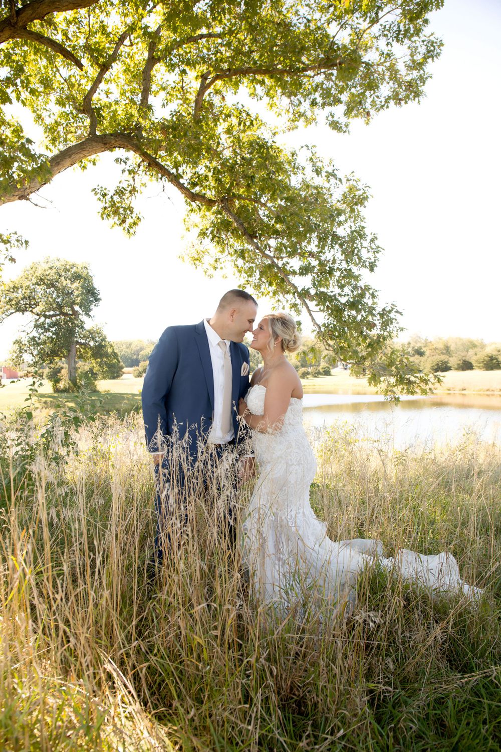 Wedding couple embraces in sunlit field with water and trees in background.
