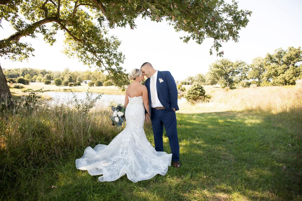 A couple shares a romantic moment under a tree during their wedding photoshoot beside a lake.