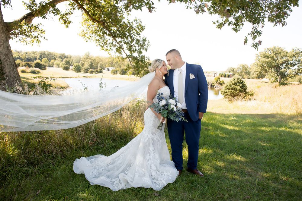 A wedding couple poses outdoors with a flowing veil against a natural landscape setting.