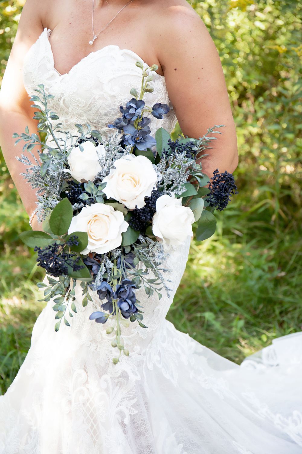 Elegant bridal bouquet featuring white roses, blue thistle, and silvery eucalyptus leaves against a lacy white wedding dress.