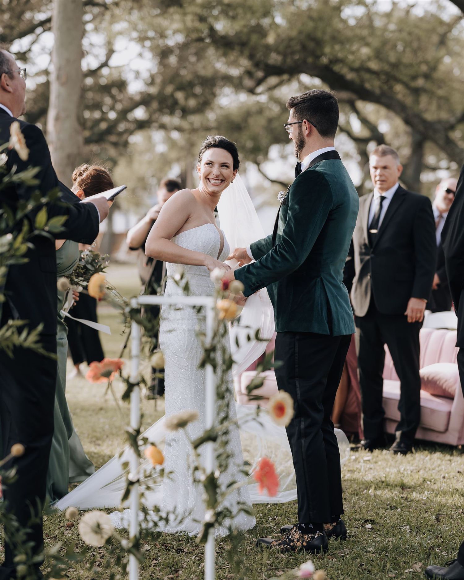 Wedding couple stands under trees during outdoor ceremony as pink flower petals float through the air.