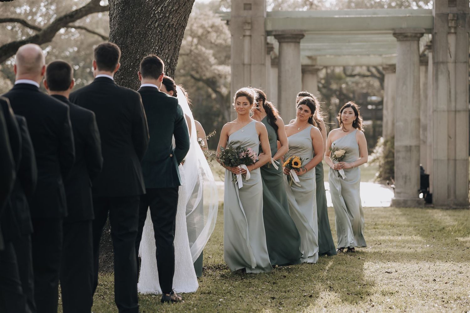 Wedding party stands in formation during outdoor ceremony with classical columns in background.