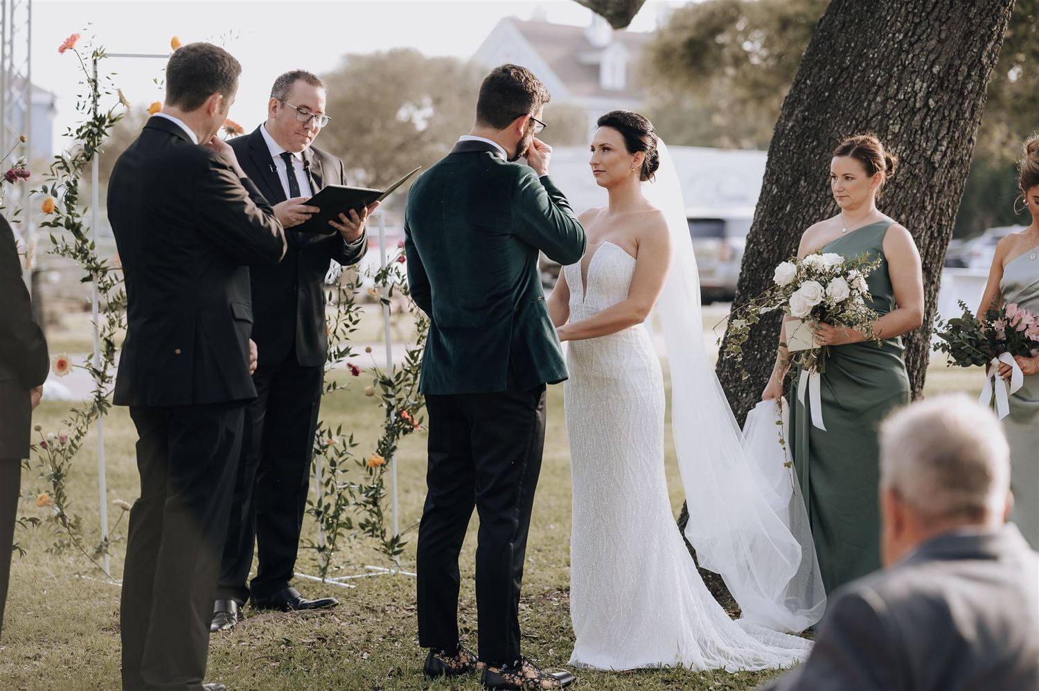 Outdoor wedding ceremony under trees with wedding party gathered at the front.