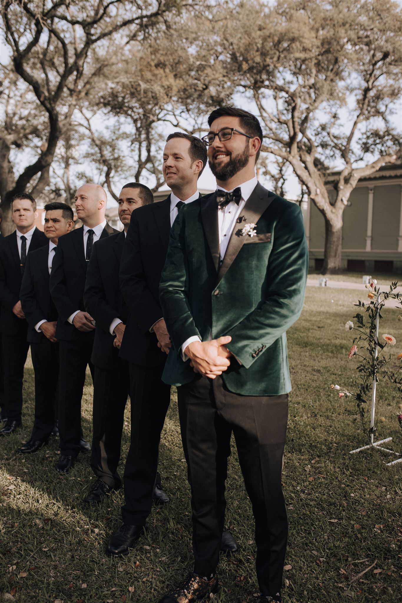 Wedding party members stand in line wearing formal attire under oak trees.