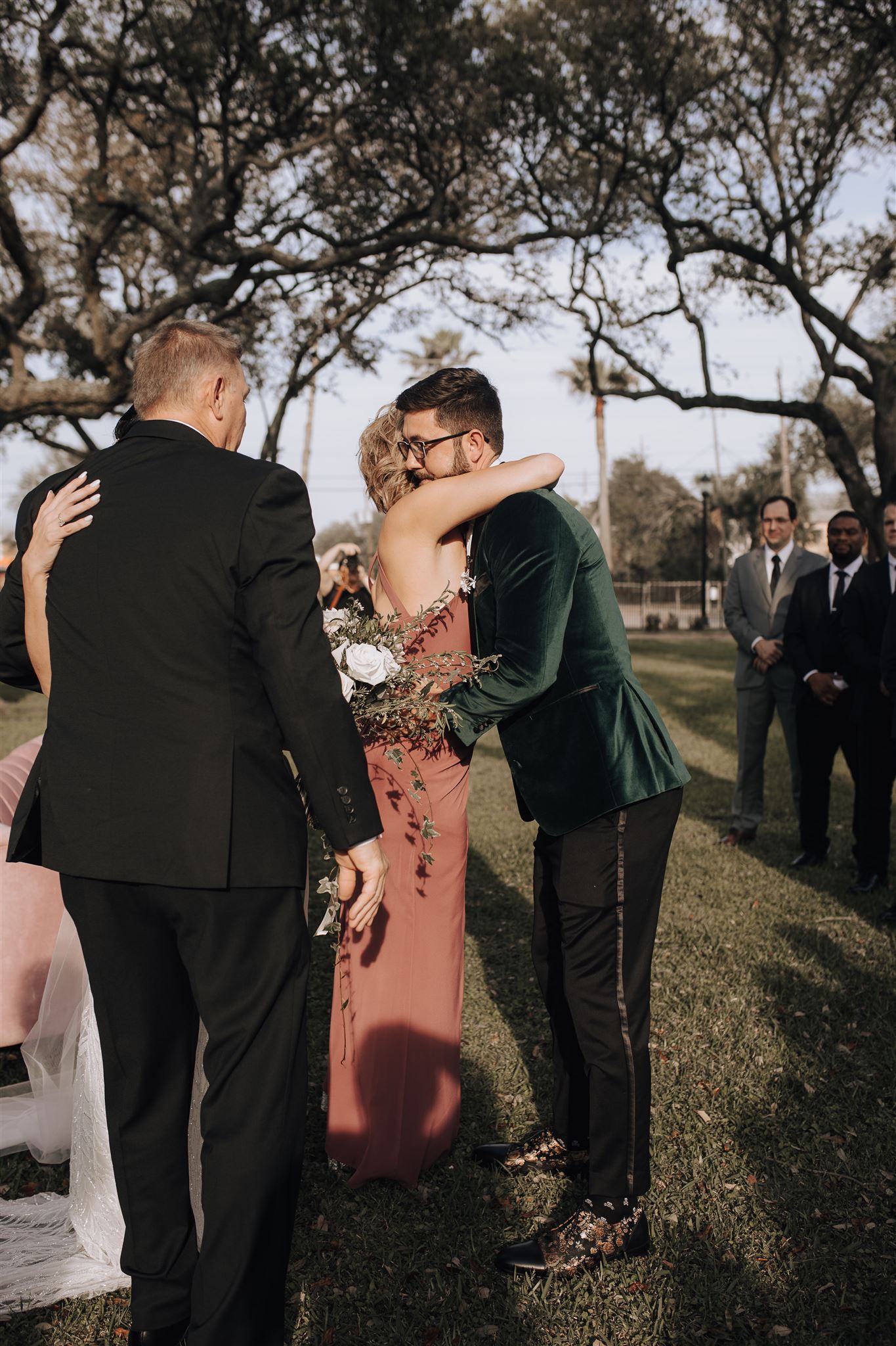 Wedding guests embrace during an outdoor ceremony under sprawling trees.