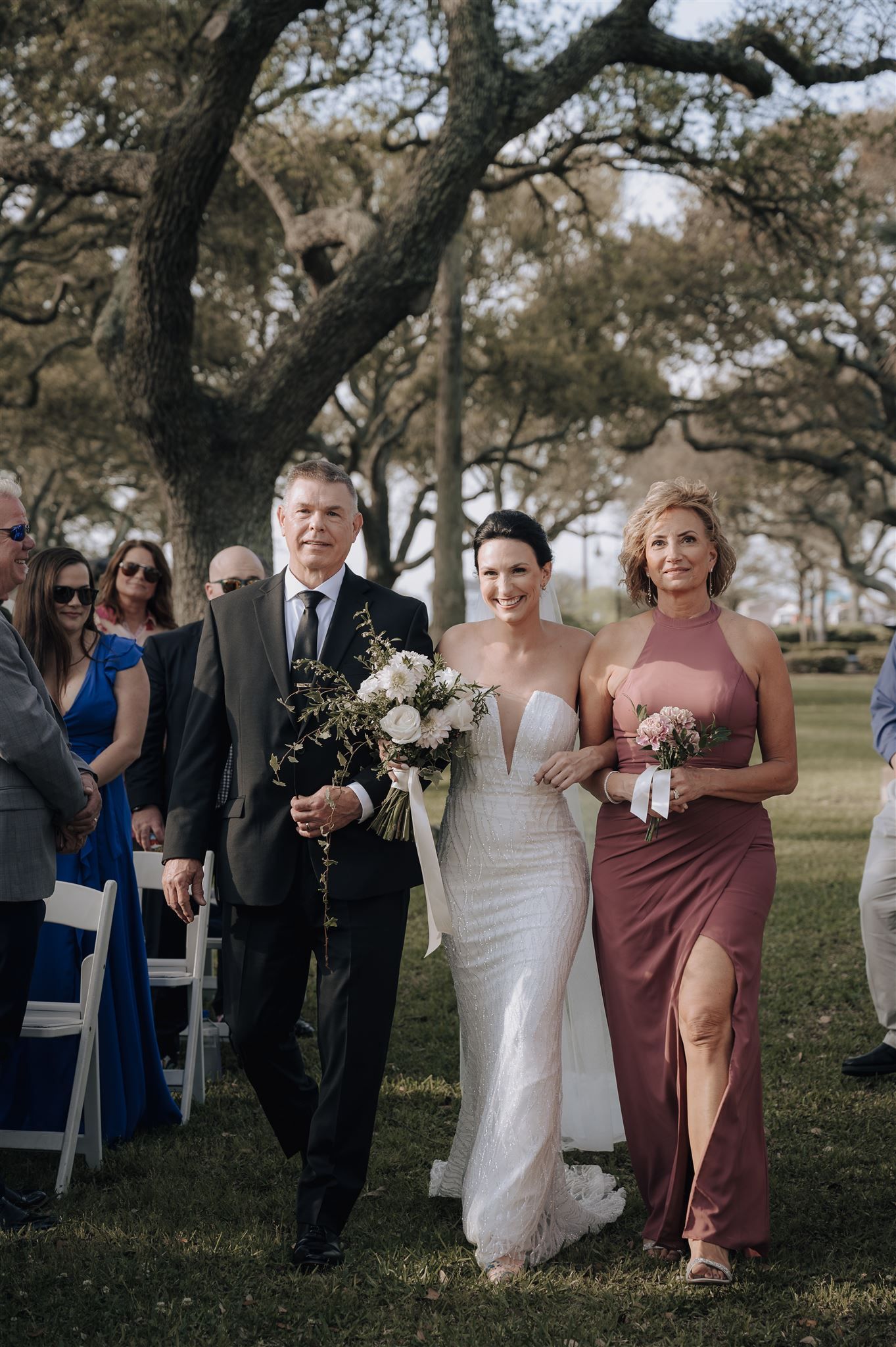 Wedding processional walks down outdoor aisle under oak trees with elegant floral bouquets.