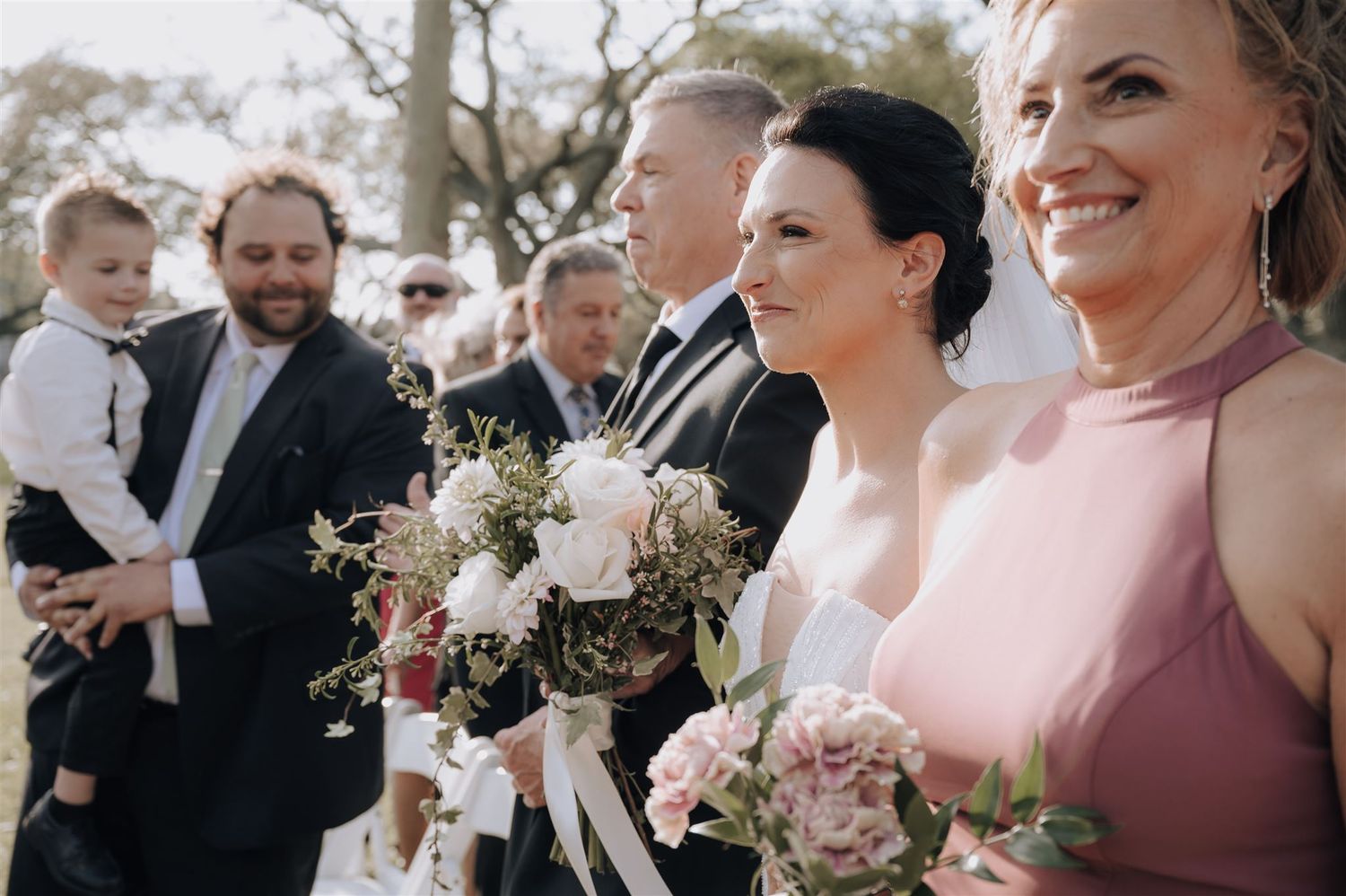 Wedding party members hold bouquets during outdoor ceremony.